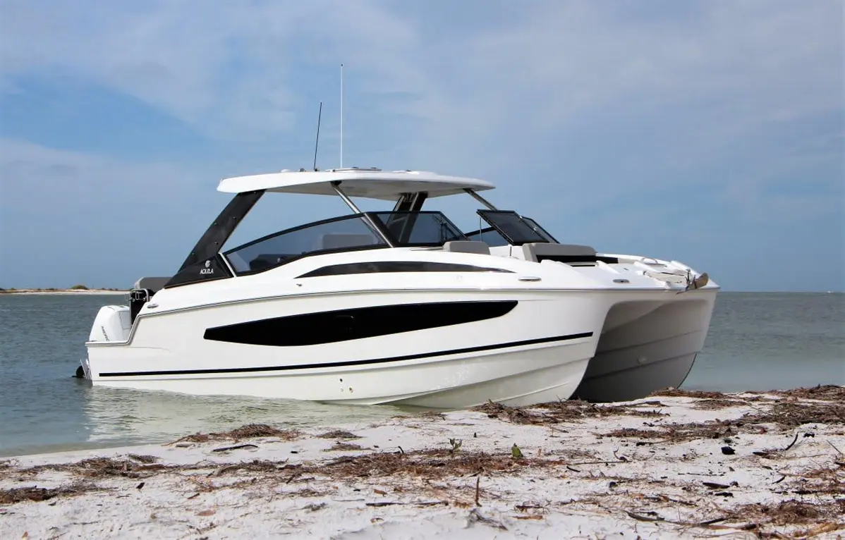 A sleek white boat with black accents is anchored on a sandy beach, with calm water and a cloudy sky in the background.